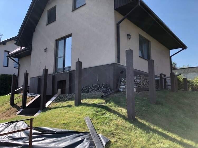 Elewacja domu jednorodzinnego z przygotowanymi słupkami pod taras, widoczne drewno opałowe i folia ochronna na trawniku.