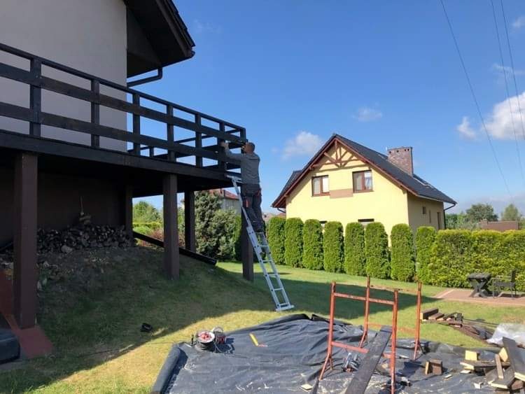Mężczyzna maluje ciemną balustradę tarasu domu jednorodzinnego, stojąc na aluminiowej drabinie, z widocznym przygotowanym miejscem pracy na trawniku.