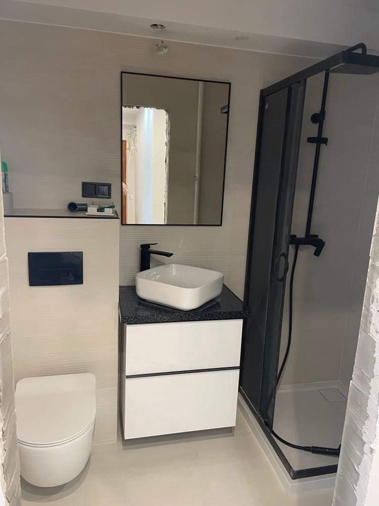 Nowoczesna łazienka z białą ceramiką, czarną armaturą i kabiną prysznicową z ciemną ramą; lustro bez ramy odbija fragment innego pomieszczenia.