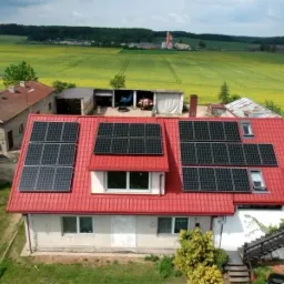 Dom jednorodzinny z czerwoną dach&oacute;wką, na kt&oacute;rej zamontowano panele fotowoltaiczne; w tle widoczne pola uprawne i zabudowania gospodarcze.
