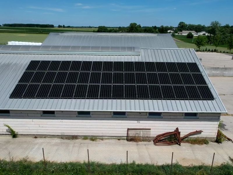 Instalacja paneli słonecznych na dachu budynku gospodarczego z blachy trapezowej, widok z góry na tle zielonych pól i błękitnego nieba.