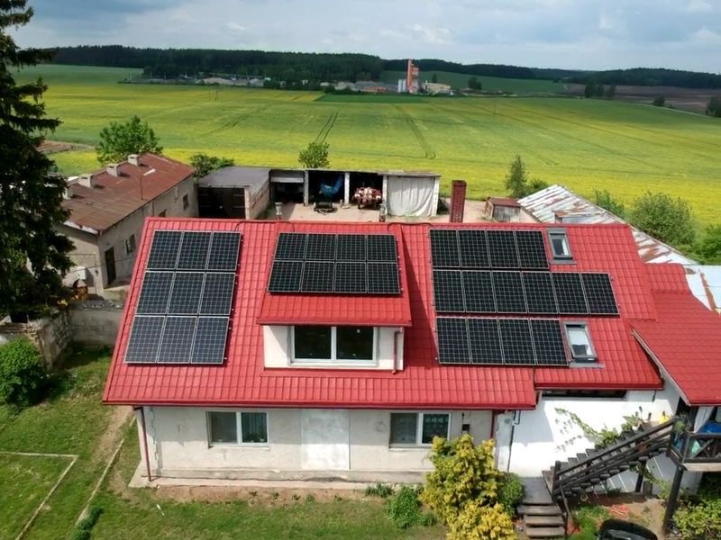 Dom jednorodzinny z czerwoną dachówką, na której zamontowano panele fotowoltaiczne; w tle widoczne pola uprawne i zabudowania gospodarcze.