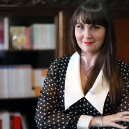 Portret kobiety w bluzce w kropki, stojącej na tle regału z książkami, z rękami skrzyżowanymi i delikatnym uśmiechem.