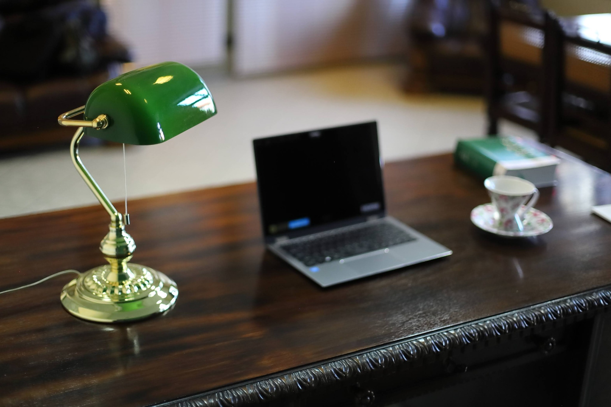 Biurko z laptopem, lampką bankierską, filiżanką i książką. Klasyczny wystrój gabinetu, ciepłe oświetlenie, ciemne drewno. Subtelne detale.