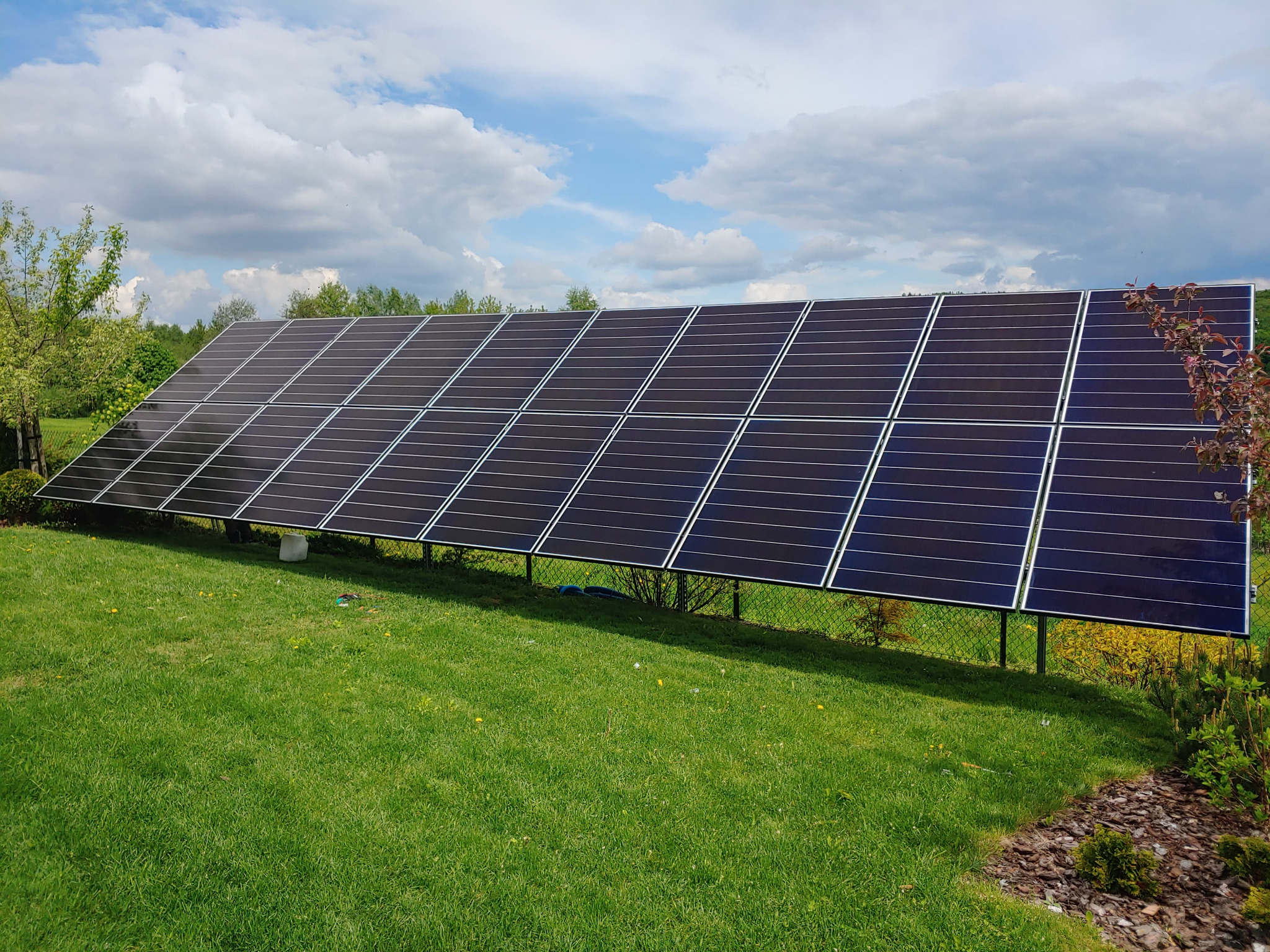 Panel słoneczny zamontowany na trawniku w ogrodzie, widoczne białe chmury na niebieskim niebie w tle.