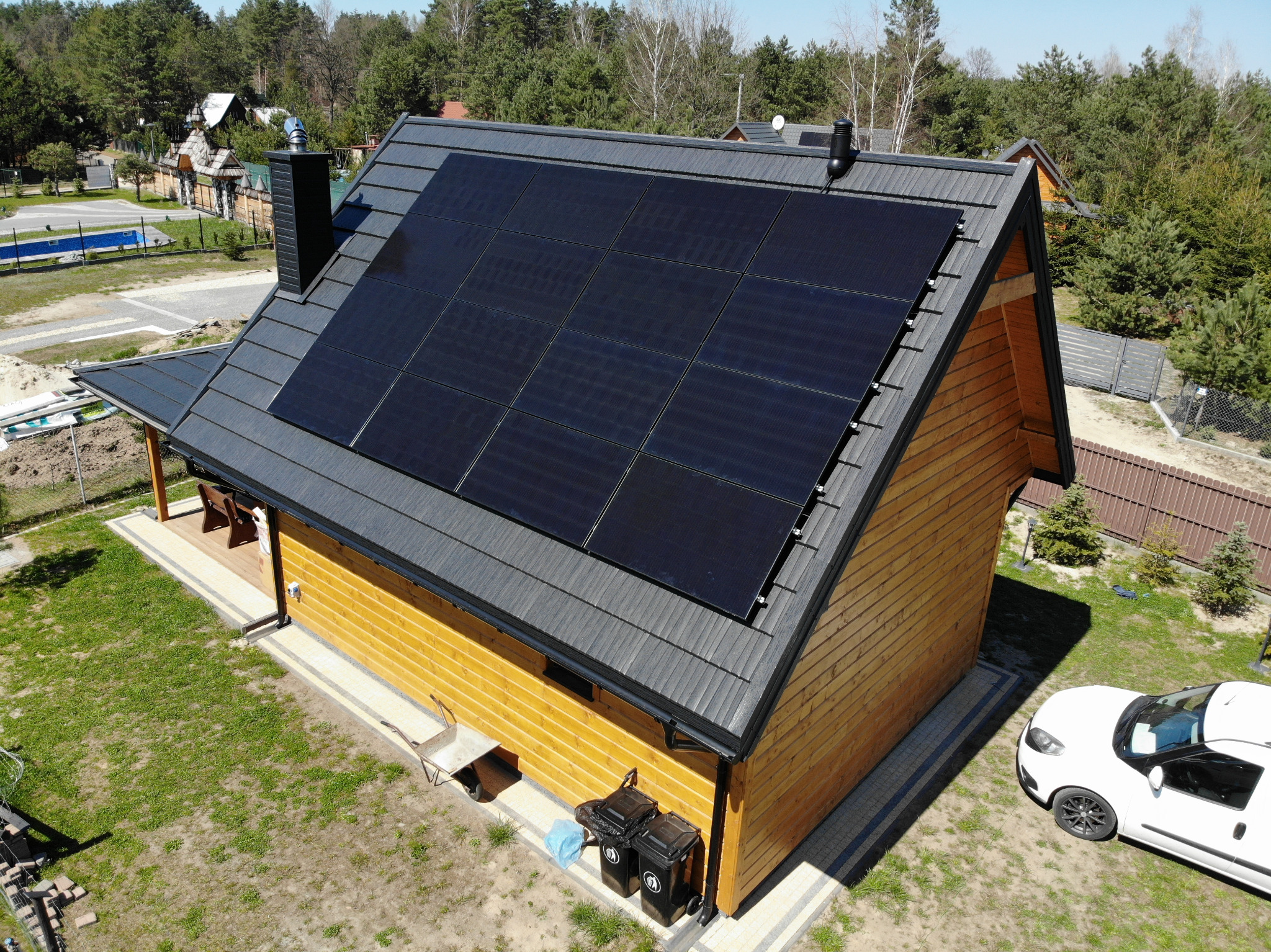 Widok z góry na dom z panelami słonecznymi na dachu, z drewnianą elewacją, czarnym dachem z blachy trapezowej, białym samochodem i dwoma koszami na śmieci obok budynku.