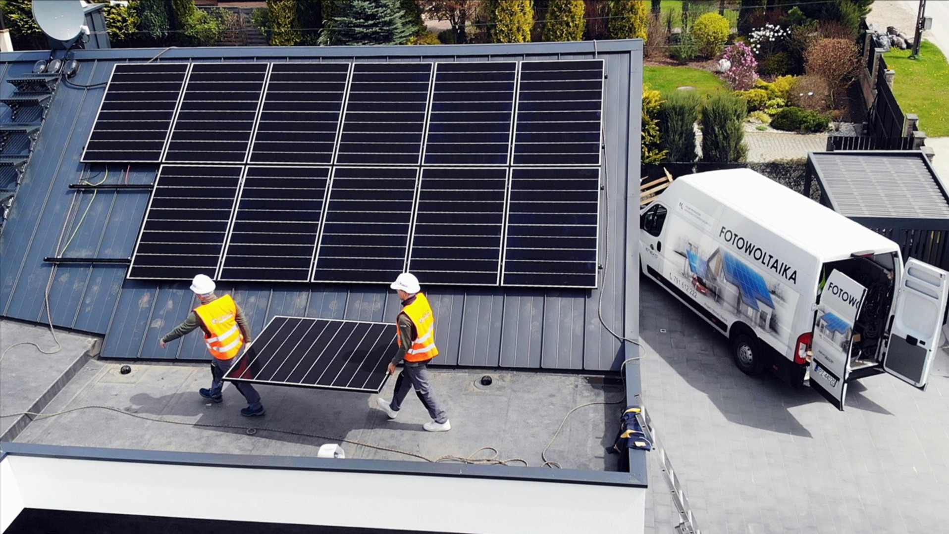 Dwóch monterów w kaskach i kamizelkach odblaskowych przenosi panel słoneczny na dachu budynku z częściowo zainstalowaną instalacją, obok zaparkowany biały samochód dostawczy z napisem 'FOTOWOLTAIKA'.