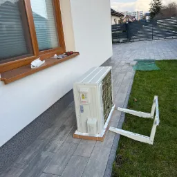 Jednostka zewnętrzna klimatyzacji, jeszcze w kartonie, przygotowana do montażu na elewacji domu, obok leżą wsporniki mocujące, na parapecie okna widoczne narzędzia.