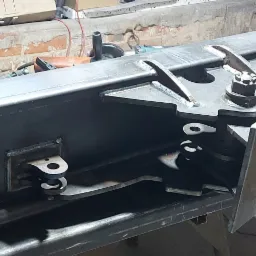 Surowy, stalowy element konstrukcyjny przypominający łyżkę koparki, spawany, z widocznymi śladami obróbki, leżący na stole warsztatowym w otoczeniu narzędzi spawalniczych, w tle ceglana ściana.