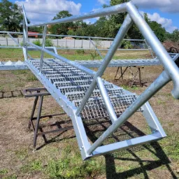 Surowe metalowe schody z kratownicowymi stopniami i poręczą, ustawione na kozłach na trawie, przygotowane do montażu zewnętrznego.