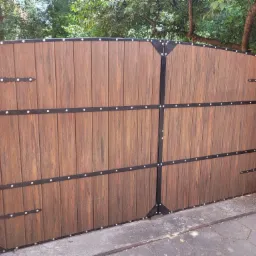 Dwuskrzydłowa brama wjazdowa z paneli imitujących drewno, z czarnymi metalowymi wzmocnieniami i dekoracyjnymi nitami, zamontowana pomiędzy ceglanymi słupkami.