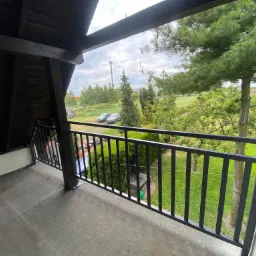 Czarna metalowa barierka balkonowa z widokiem na zielony ogród, samochody i wieżę transmisyjną w tle.