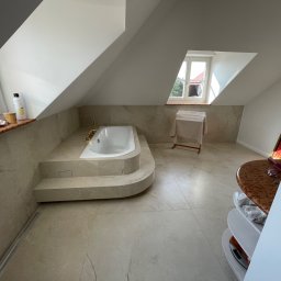 LUK Łukasz Zatorski - Jasna łazienka na poddaszu z wanną zabudowaną w marmurowym podeście, widok na okno dachowe i fragment półki z kosmetykami.