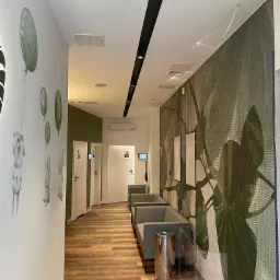 Korytarz w Legionowie z drewnopodobną podłogą, ścianą z motywem roślinnym i rysunkami zwierząt z balonami, szara poczekalnia z koszem na śmieci ze stali nierdzewnej.