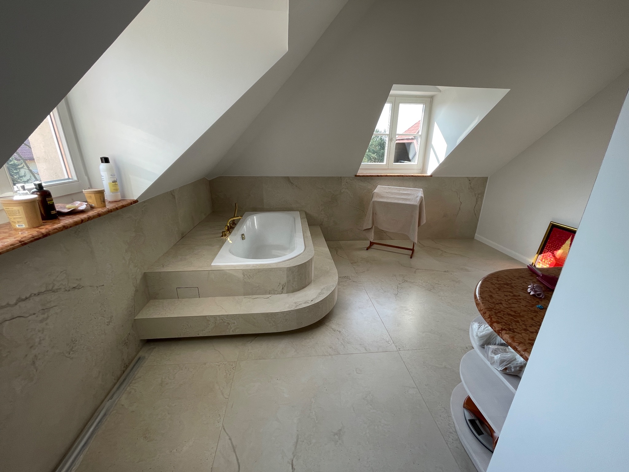 Jasna łazienka na poddaszu z wanną zabudowaną w marmurowym podeście, widok na okno dachowe i fragment półki z kosmetykami.