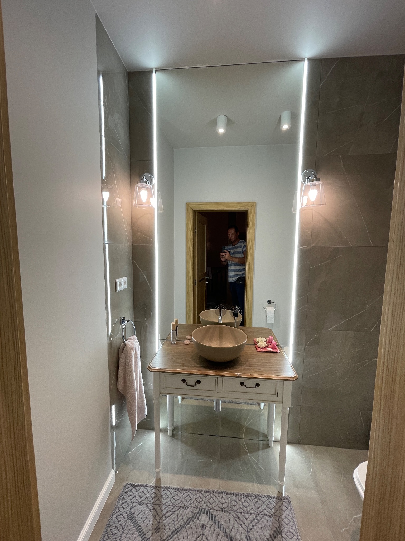 Nowoczesna łazienka z umywalką nablatową w odcieniach beżu, podświetlanym lustrem i szarymi płytkami imitującymi marmur, widoczny fragment dywanika i drzwi.