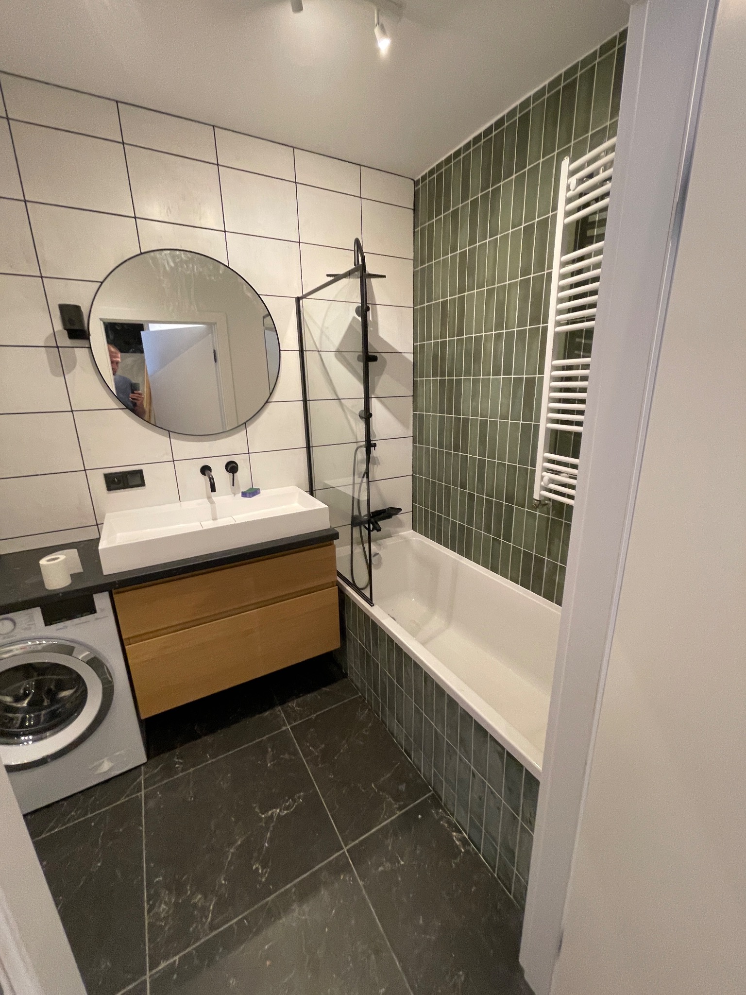 Nowoczesna łazienka z białymi płytkami, okrągłym lustrem, umywalką nablatową na drewnianej szafce, pralką i wanną obłożoną zielonymi płytkami.