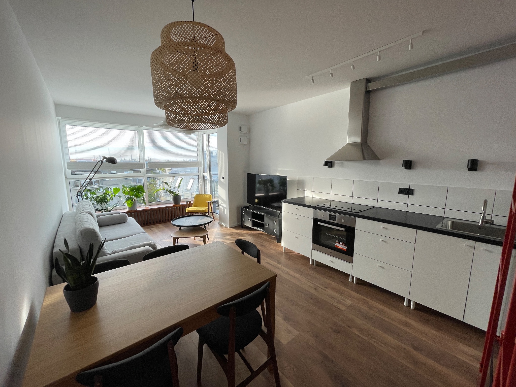 Jasne, przestronne wnętrze mieszkania z otwartą kuchnią, drewnianym stołem jadalnym i sofą przy oknie, oświetlone wiszącą lampą z naturalnego materiału.