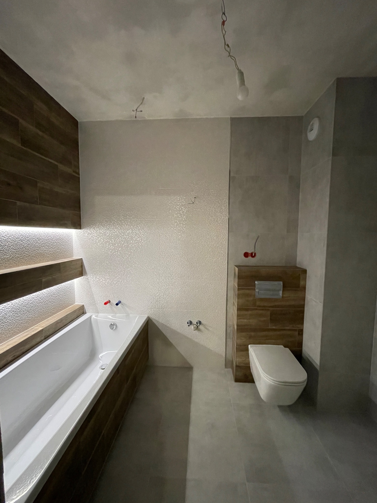 Nowoczesna łazienka po remoncie: biała wanna z drewnianym obramowaniem i podświetleniem LED, toaleta podwieszana z obudową w drewnie, szare płytki podłogowe i ścienne, surowy betonowy sufit...