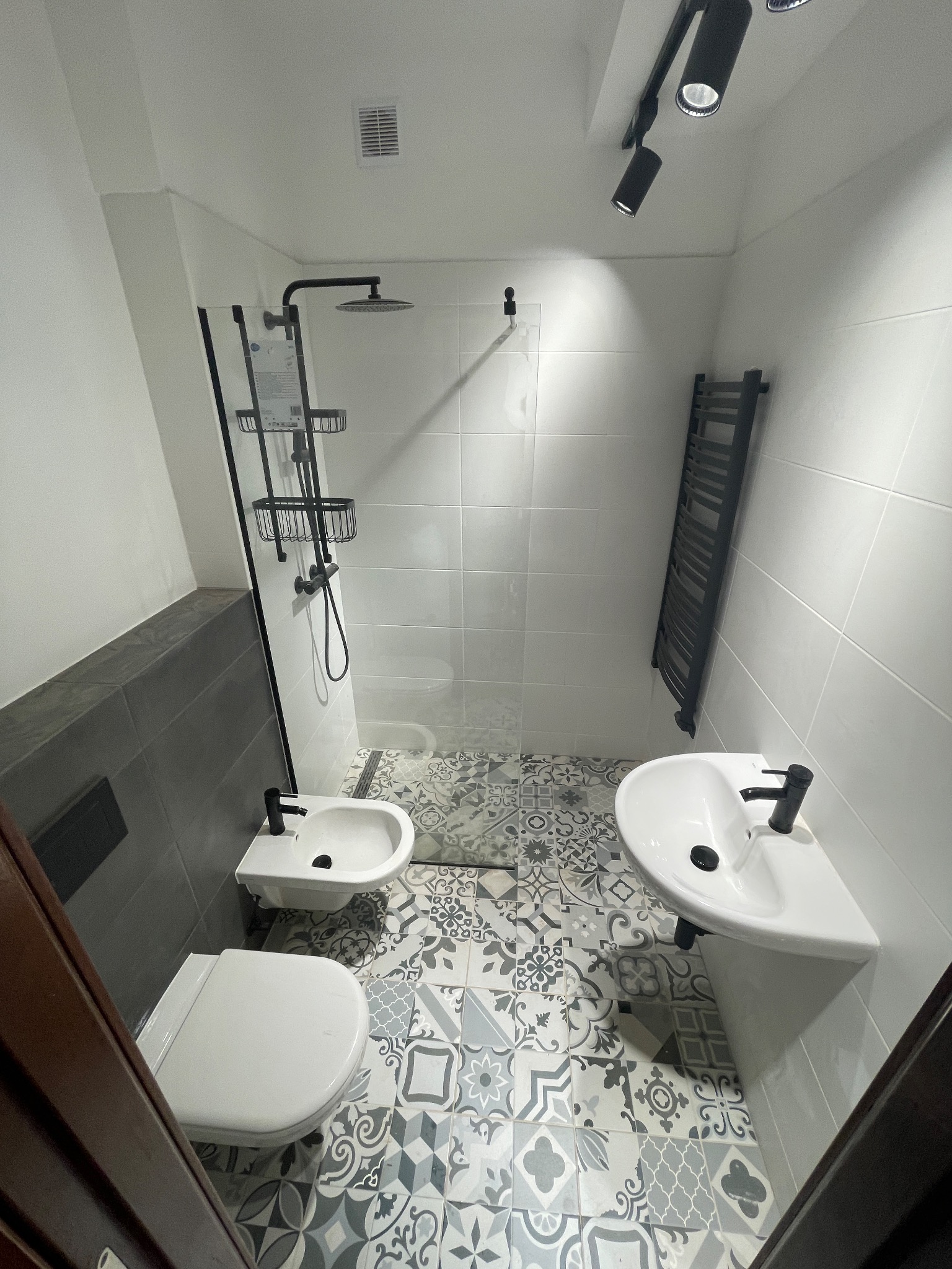 Nowoczesna łazienka z kabiną prysznicową typu walk-in, umywalką na postumencie i bidetem, wykończona płytkami podłogowymi w stylu patchwork oraz szarymi płytkami częściowo na ścianach.