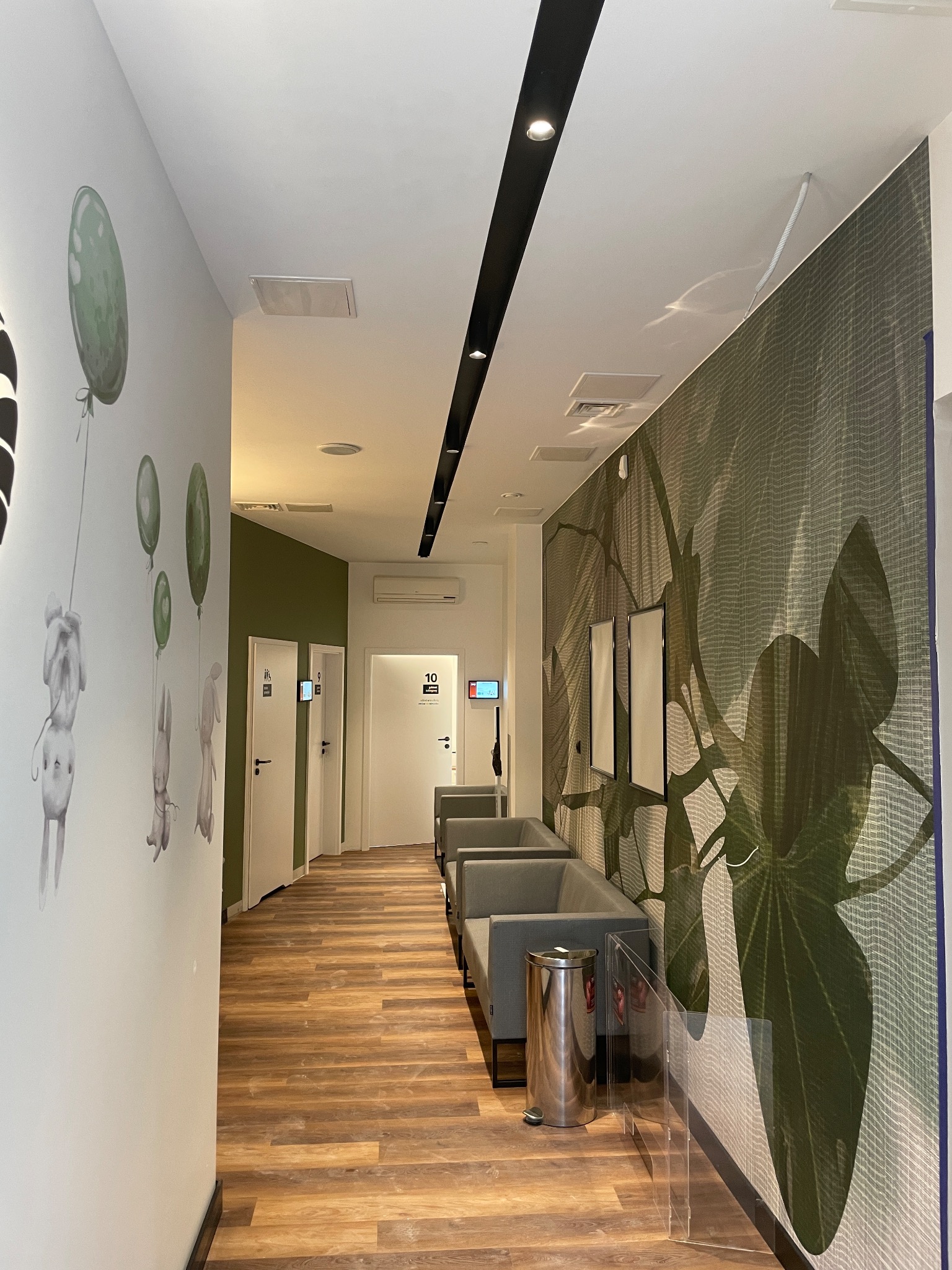 Korytarz w Legionowie z drewnopodobną podłogą, ścianą z motywem roślinnym i rysunkami zwierząt z balonami, szara poczekalnia z koszem na śmieci ze stali nierdzewnej.
