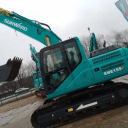 Turkusowa koparka Sunward SWE155F na placu, z podniesionym ramieniem i łyżką, widoczne inne koparki w tle oraz flaga producenta.