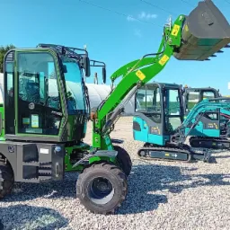 Zielona ładowarka kołowa z podniesioną łyżką na tle innych maszyn budowlanych i błękitnego nieba.