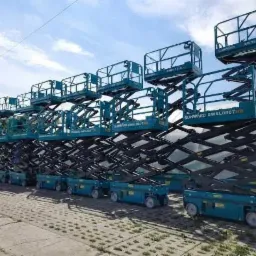 Rząd turkusowych podnośników nożycowych Sunward SWSL0807HD ustawionych na zewnątrz na brukowanym placu, widoczne czarne mechanizmy nożycowe i niebieskie platformy robocze z barierkami, błękitne...