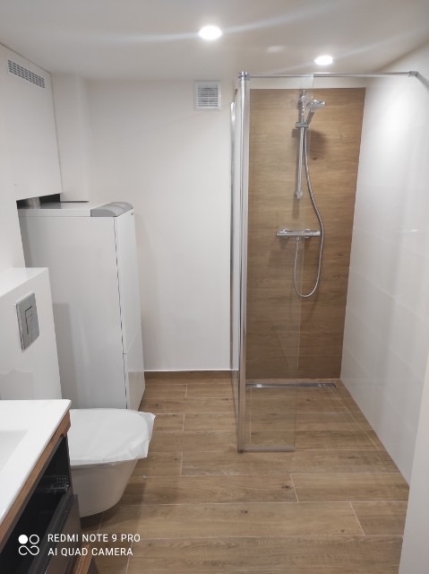 Nowoczesna łazienka z kabiną prysznicową typu walk-in, wykończona płytkami imitującymi drewno, z białą zabudową i toaletą wiszącą, oświetlona punktowo.