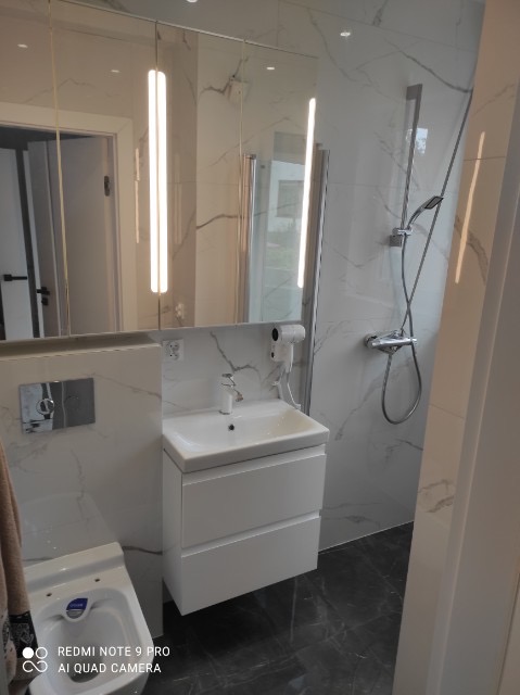 Nowoczesna łazienka z białą umywalką nablatową, szafką pod umywalką, bidetem i kabiną prysznicową wykończona płytkami imitującymi marmur.