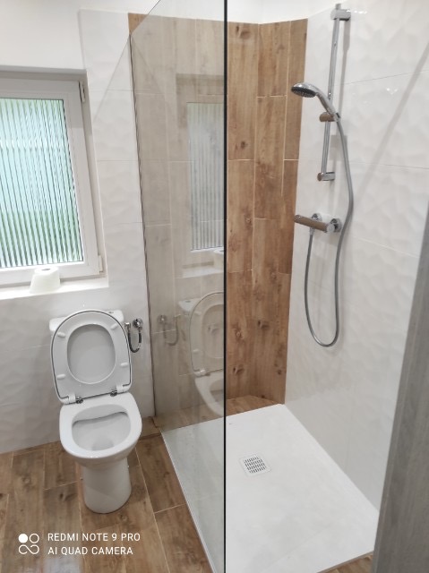 Nowoczesna łazienka z kabiną prysznicową typu walk-in oddzieloną szklaną ścianką, białą armaturą i panelami imitującymi drewno na ścianie przy prysznicu.