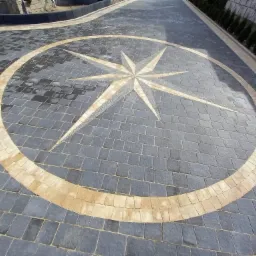 Ułożona kostka brukowa w szarym kolorze z dekoracyjnym wzorem rozety w odcieniach beżu i szarości, tworząca unikalny wz&oacute;r na podjeździe.