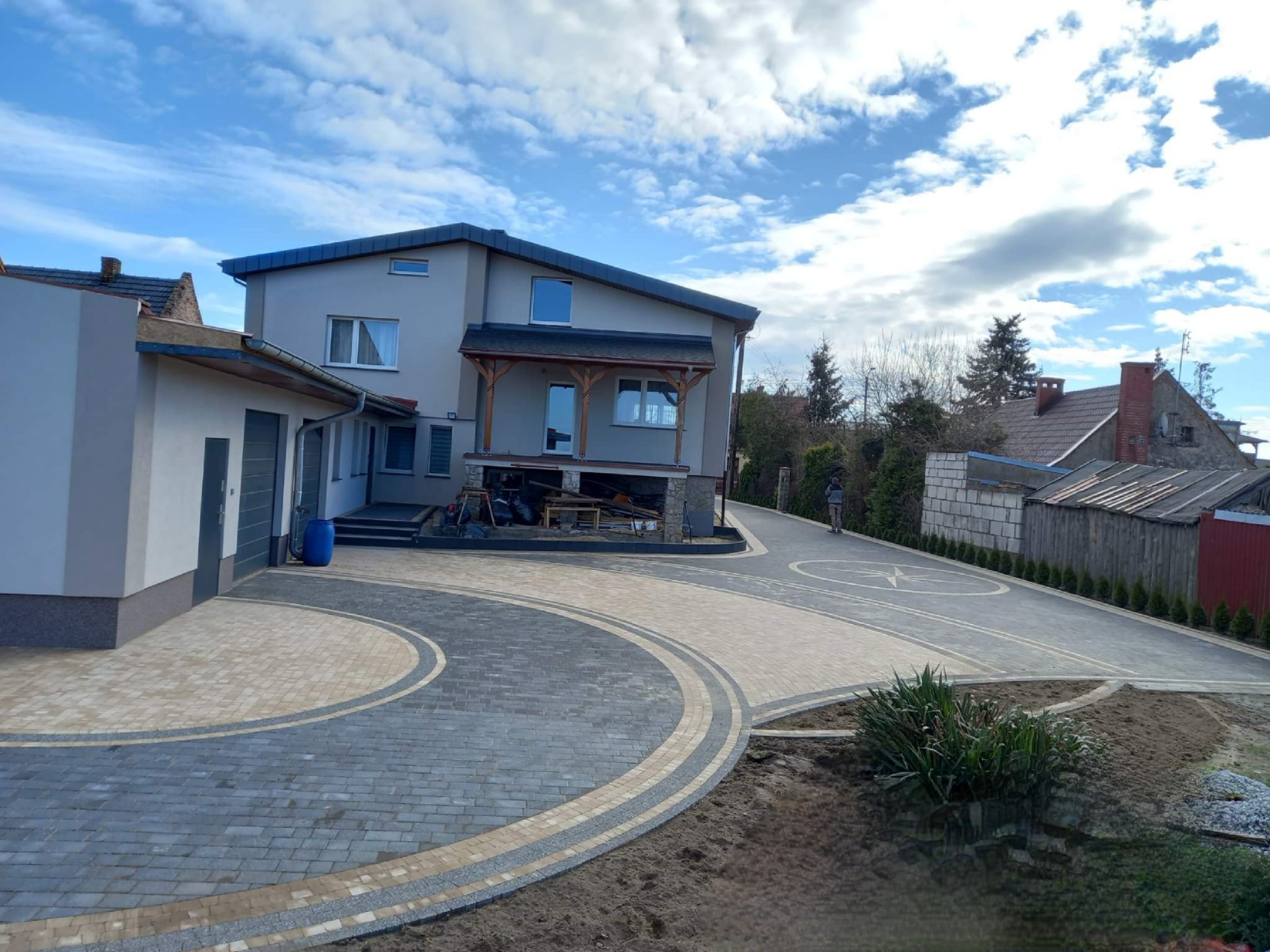 Nowoczesny podjazd z kostki brukowej w odcieniach szarości i beżu, ułożony w dekoracyjny wzór przed domem z drewnianym gankiem, błękitne niebo z chmurami w tle.