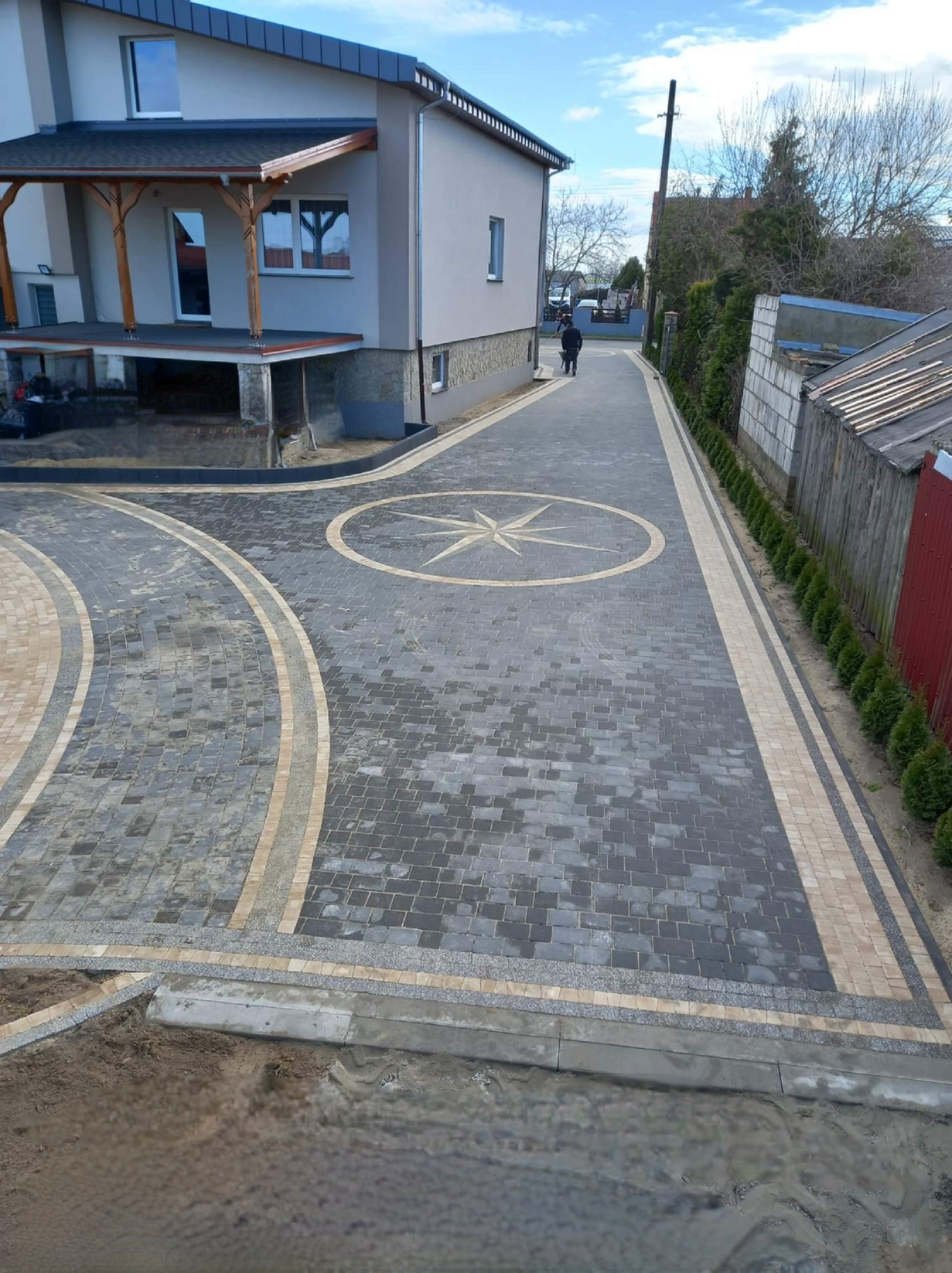 Ułożona z kostki brukowej aleja prowadząca do domu, z dekoracyjnym motywem rozety pośrodku i obrzeżami z jaśniejszej kostki, otoczona tujami i ogrodzeniem.