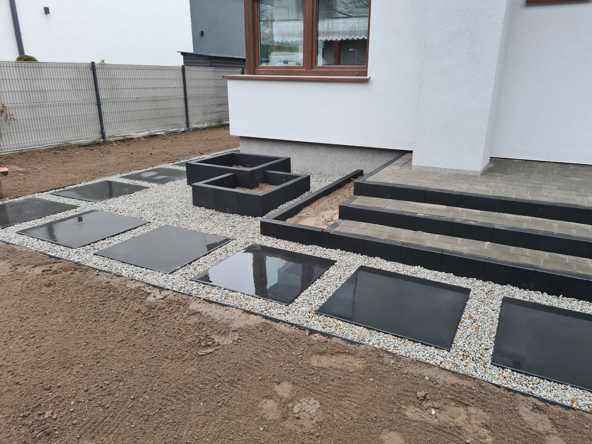 Nowoczesna aranżacja ogrodu z ciemnymi, kwadratowymi płytami chodnikowymi ułożonymi na jasnym żwirze, prowadzącymi do schodów wejściowych i podwyższonych rabat z czarnej kostki.