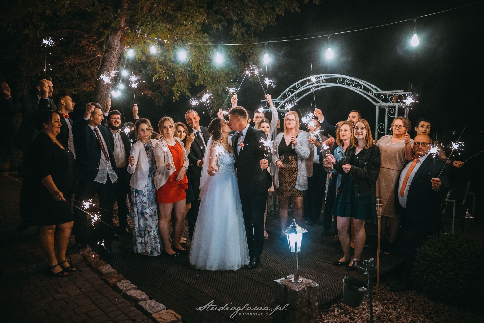 Pocałunek pary młodej otoczonej gośćmi z zimnymi ogniami, wieczorowa sceneria z dekoracyjnymi lampkami i białą altaną w tle.