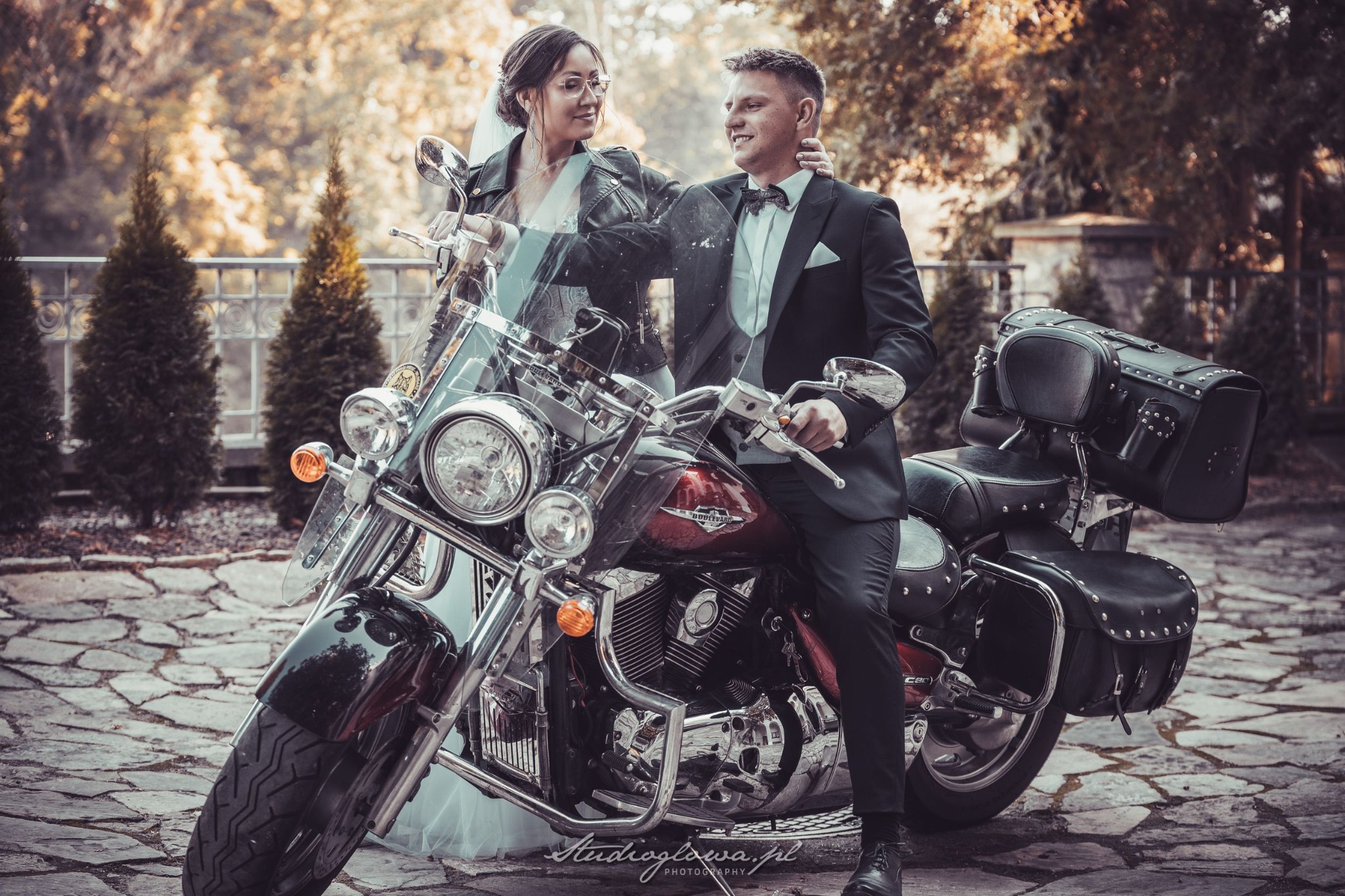 Para młoda w strojach ślubnych pozuje na motocyklu typu cruiser na tle parkowej scenerii z tujami i kamienną nawierzchnią.