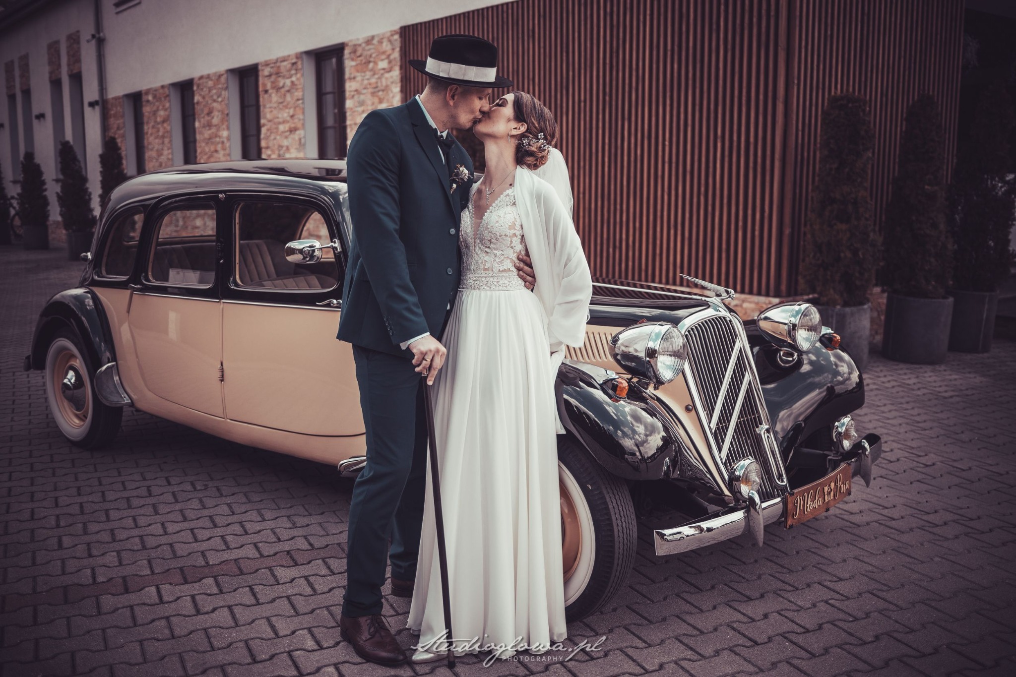 Pocałunek nowożeńców w stylu retro przed klasycznym samochodem z drewnianą tabliczką 'Młoda Para'. Panna młoda w koronkowej sukni i białym szalu, pan młody w zielonym garniturze i kapeluszu.