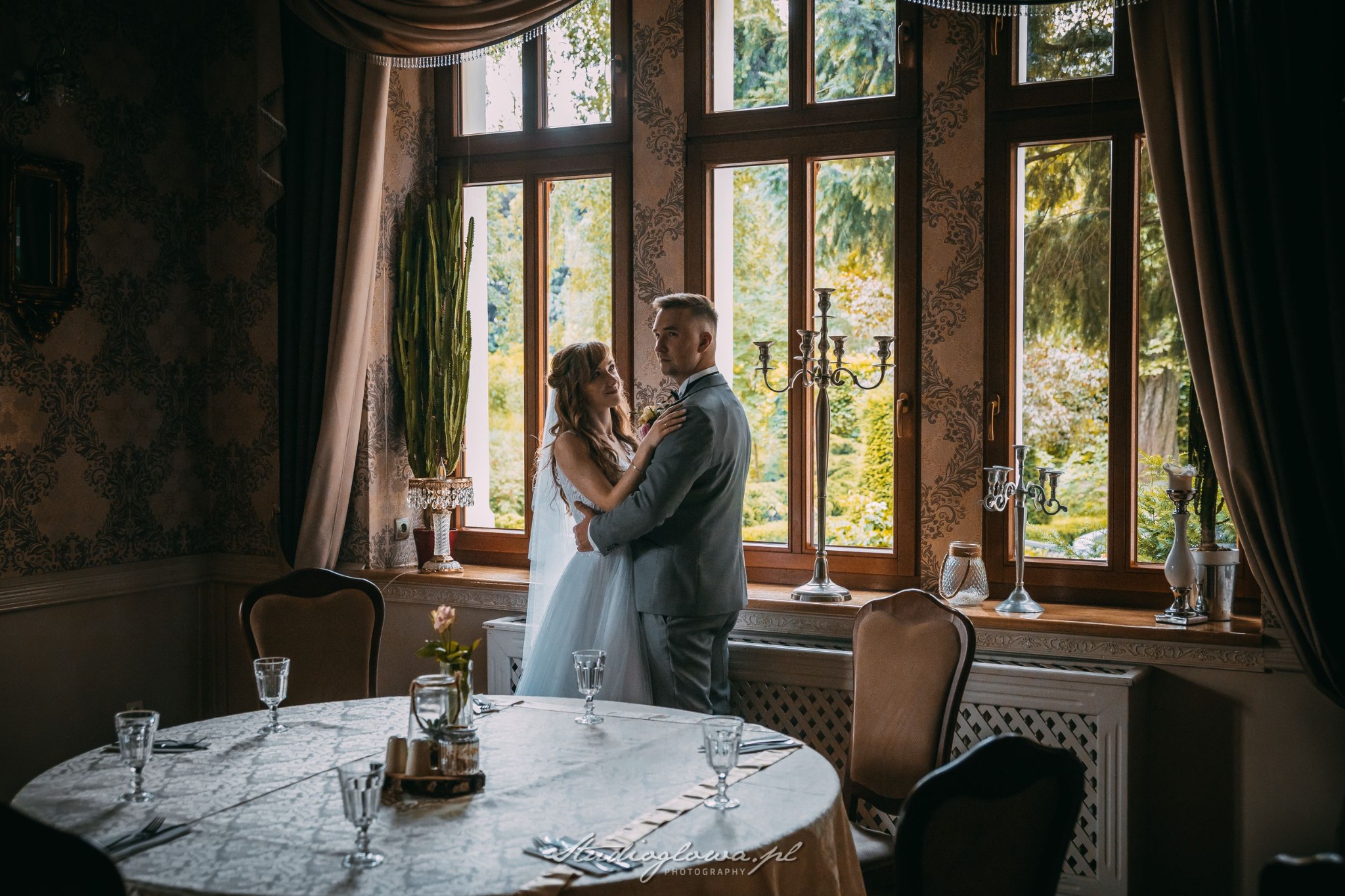 Para młoda w objęciach na tle okna w stylowym wnętrzu pałacu, widoczny stół z nakryciem i dekoracjami.