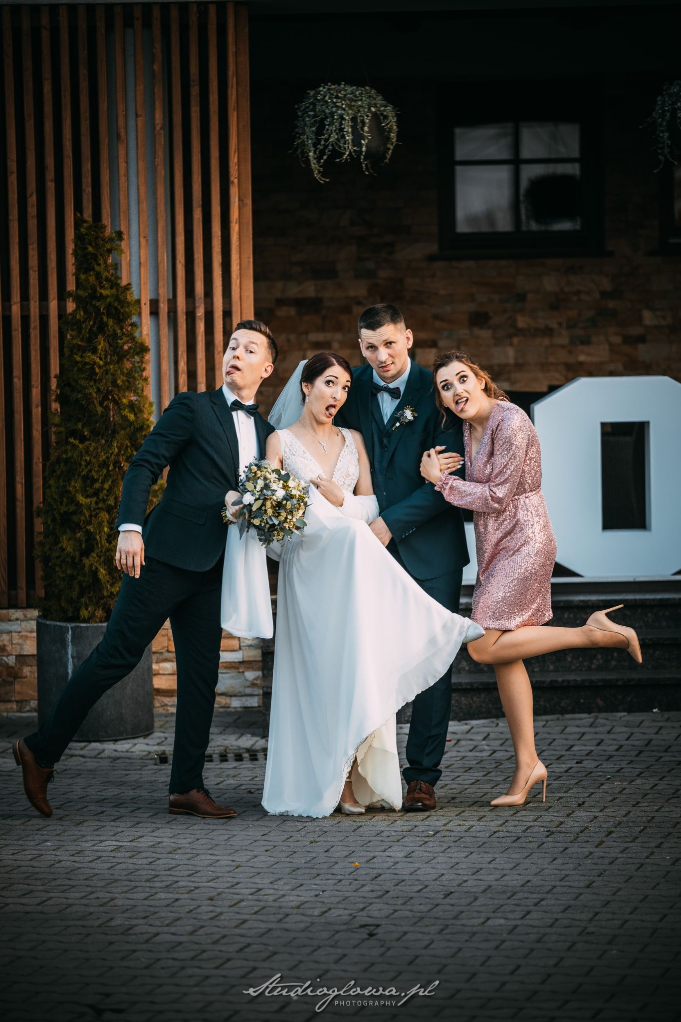 Panna młoda w białej sukni i pan młody w granatowym garniturze pozują z gośćmi weselnymi, robiąc śmieszne miny na tle dekoracji z liter 'LOVE'.