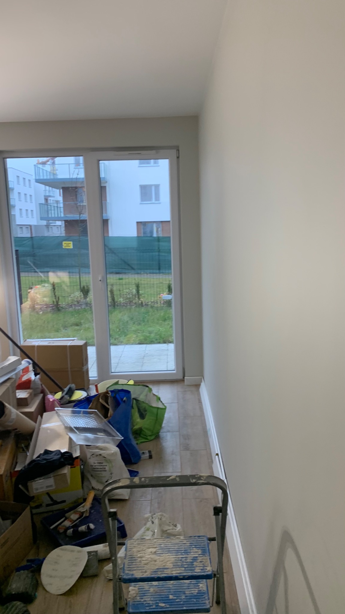 Przygotowania do malowania w pokoju: drabina malarska, wiadro z farbą, pędzle i inne akcesoria malarskie zgromadzone przy ścianie i oknie balkonowym, widoczne nowe budynki za oknem.