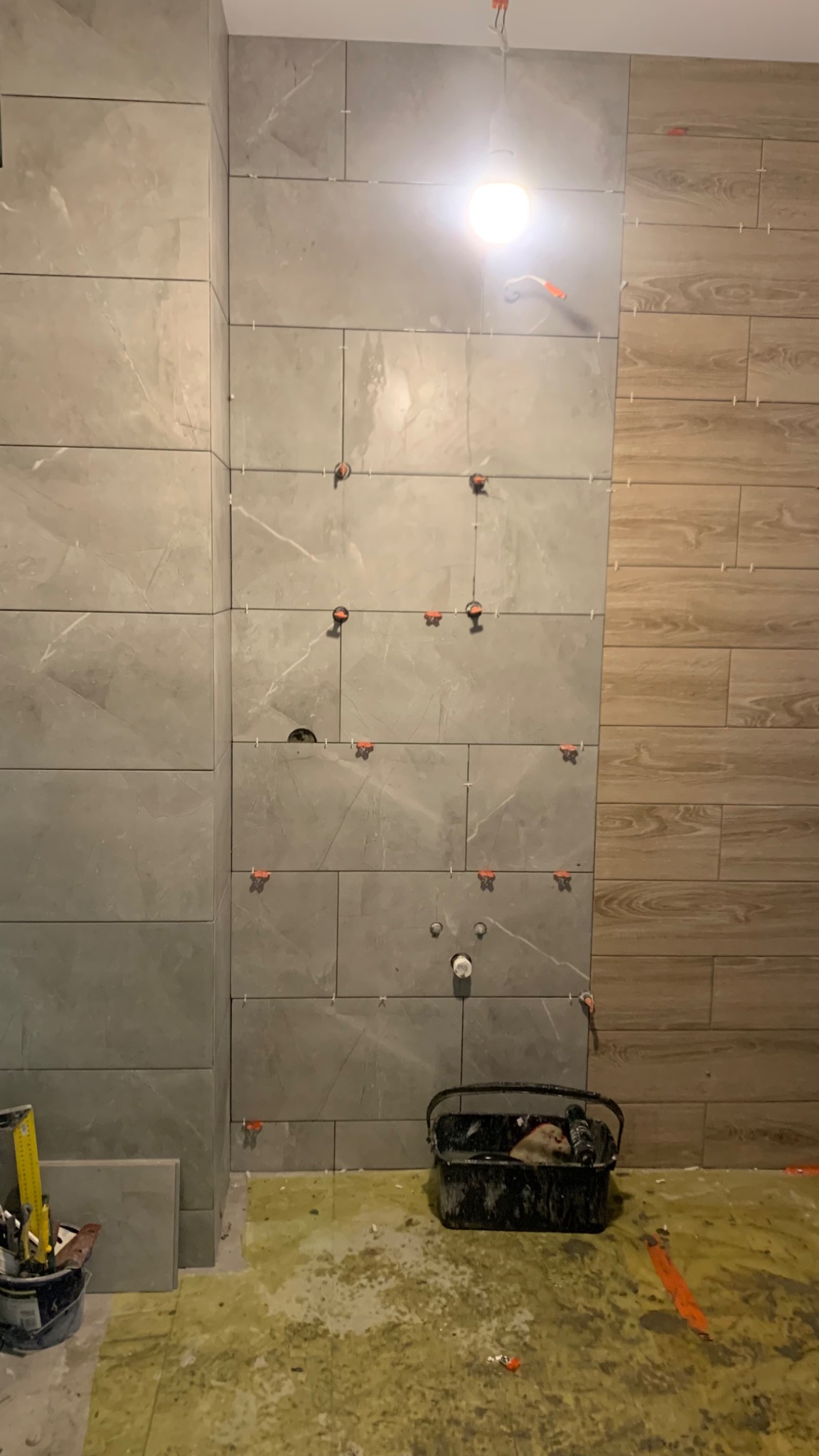 Ściana częściowo wyłożona szarymi płytkami imitującymi marmur, z widocznymi krzyżykami dystansowymi i systemem poziomowania płytek, obok ściana wykończona panelami drewnopodobnymi, na podłodze...