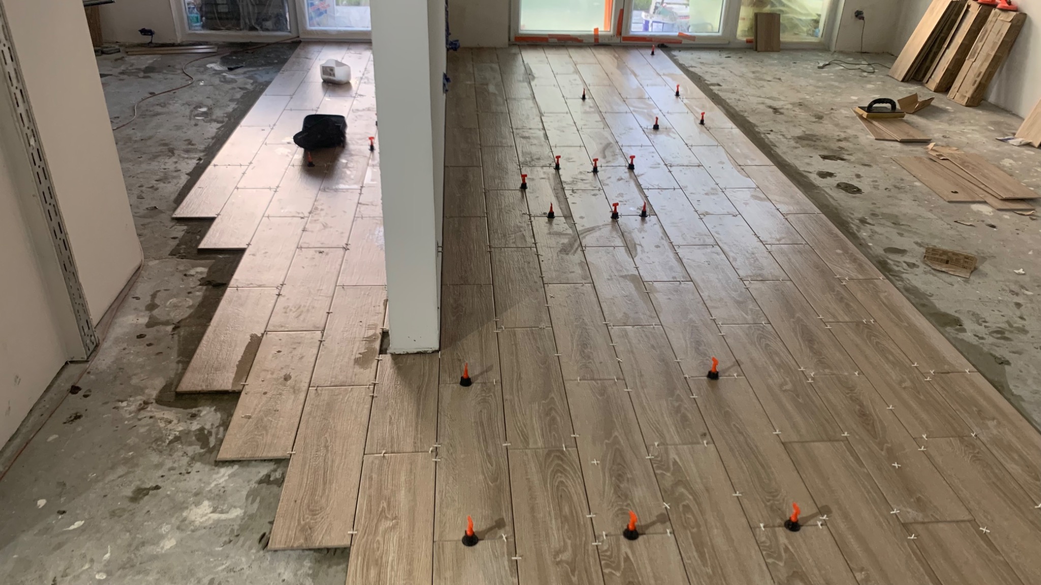 Układanie podłogi z płytek imitujących drewno, z użyciem poziomujących klipsów i klinów, widoczne narzędzia glazurnicze i niezakończone prace w pomieszczeniu z oknami.