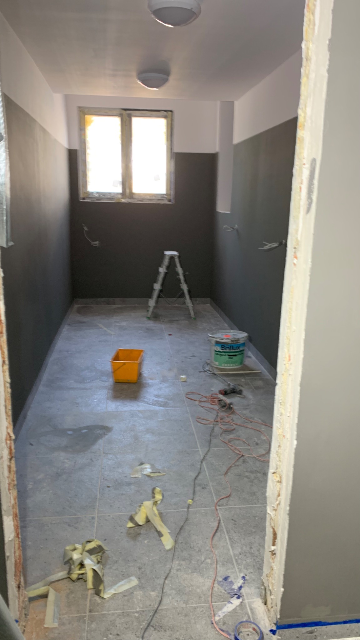 Pokój w trakcie malowania: ściany pomalowane do połowy na szaro, drabina, wiadro z farbą, kuweta i kable na podłodze.