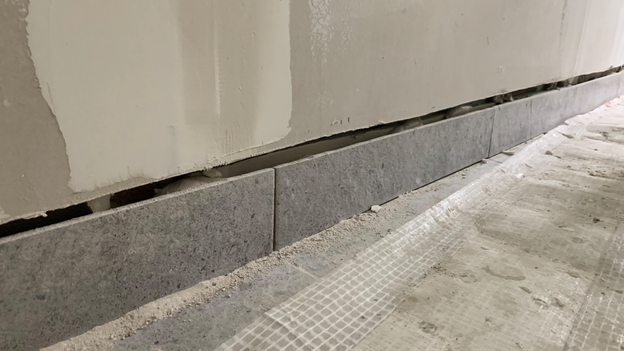 Szara, kamiennopodobna listwa przypodłogowa, częściowo zamontowana na ścianie ze śladami szpachli, z widocznym odstępem od ściany i zabezpieczoną podłogą.