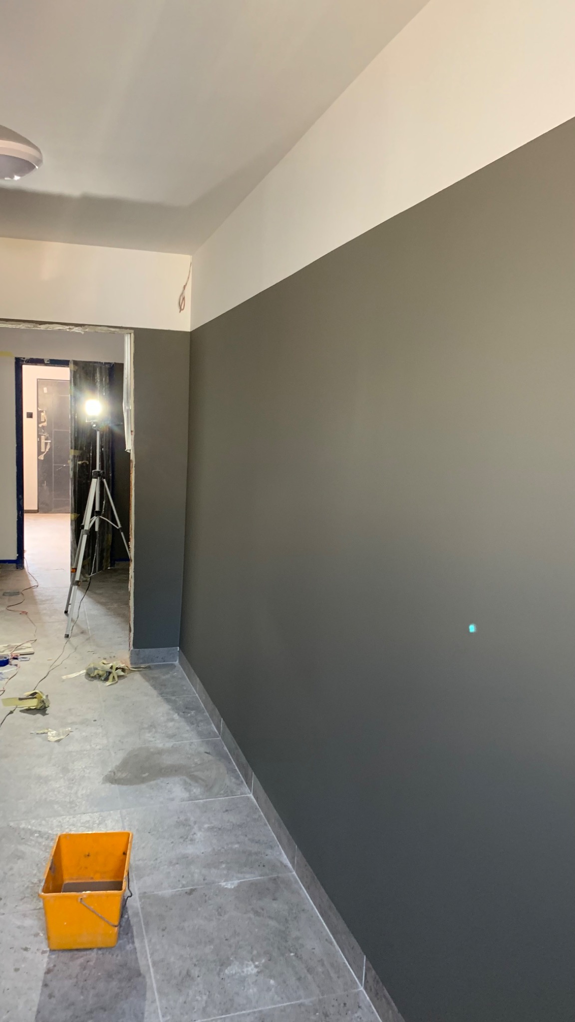 Świeżo pomalowana szara ściana z białym pasem u góry, widoczny żółty kubeł na podłodze i lampa na statywie w przejściu do innego pomieszczenia.