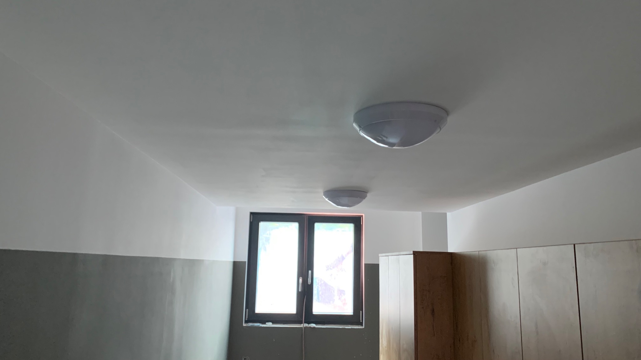 Pomieszczenie po odświeżeniu ścian, z szarą lamperią i białymi górnymi partiami, widoczne dwie okrągłe lampy sufitowe i fragmenty szaf.