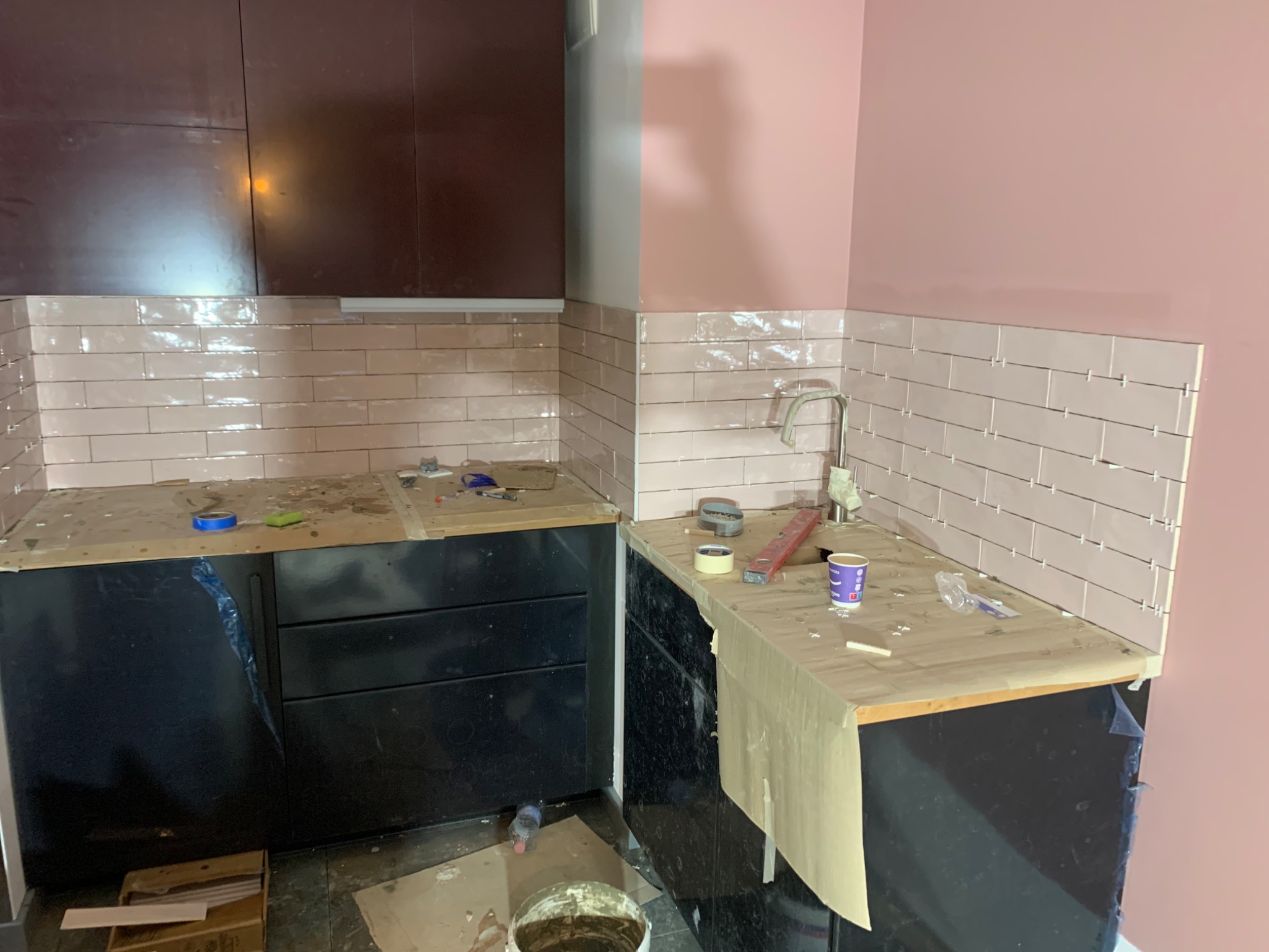 Układanie jasnoróżowych płytek w kuchni w Rumii, widoczna część ściany z nowymi płytkami, czarne szafki kuchenne z zabezpieczonym blatem, wiadro z zaprawą i narzędzia glazurnicze.