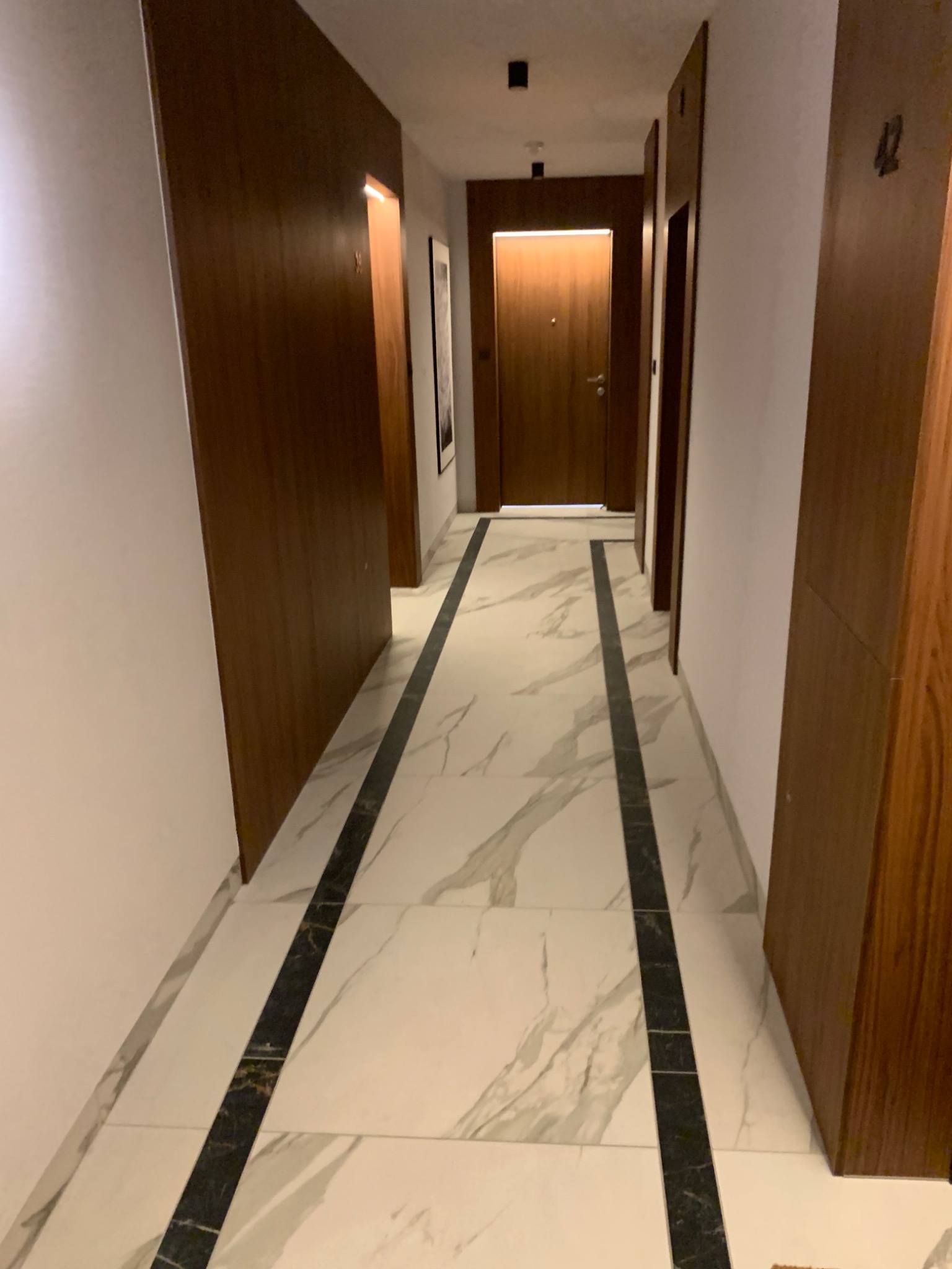 Korytarz wykończony białymi płytkami z szarymi żyłami i czarnymi pasami, z ciemnymi drewnianymi drzwiami po obu stronach.
