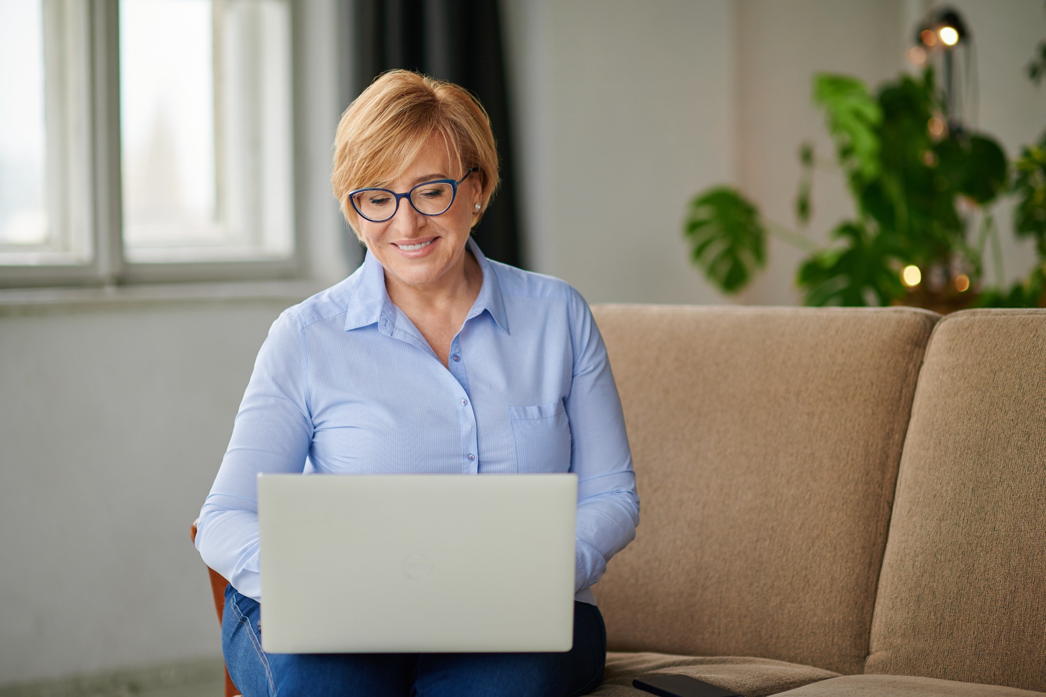 Uśmiechnięta kobieta w okularach, siedząca na beżowej kanapie, spogląda w dół na otwarty laptop.